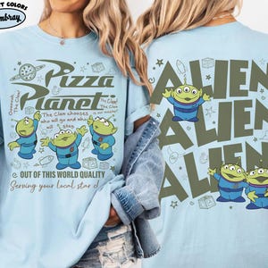 May include: Light blue t-shirt featuring a Pizza Planet design with three-eyed green aliens. The front of the shirt has the text "Pizza Planet" and "Out of this world quality". The back of the shirt has the word "ALIEN" repeated.