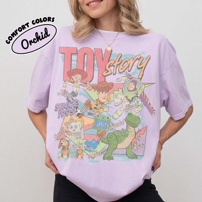 May include: A light purple t-shirt featuring a graphic print of Toy Story characters including Woody, Buzz Lightyear, Jessie, Rex, and Slinky Dog. The text reads Toy Story, Since 1995, Jessie, Woody, Buzz, Smarty Pants, Hi! Lilypad, Atlas, and Slinky.