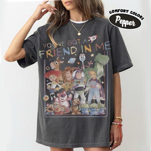 May include: A charcoal gray short-sleeve t-shirt featuring a graphic print of various animated characters from the Toy Story franchise. The text "YOU'VE GOT A FRIEND IN ME" is printed above the characters. A "COMFORT COLORS Pepper" label is visible.