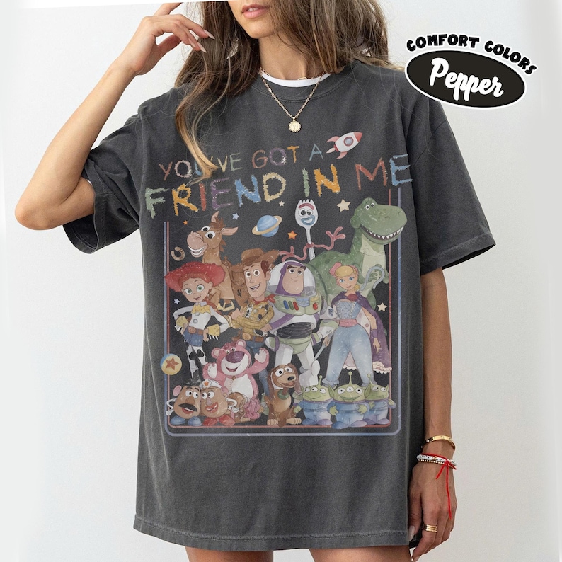 May include: A charcoal gray short-sleeve t-shirt featuring a graphic print of various animated characters from the Toy Story franchise. The text "YOU'VE GOT A FRIEND IN ME" is printed above the characters. A "COMFORT COLORS Pepper" label is visible.