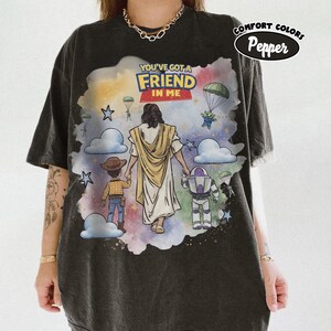 May include: Dark gray t-shirt with a colorful graphic featuring the text "YOU'VE GOT A FRIEND IN ME". The design includes cartoon characters and a figure in a white robe. The shirt has a relaxed fit and short sleeves.