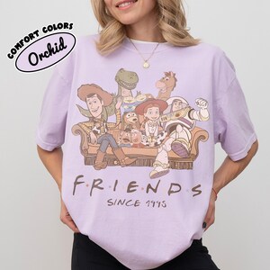 May include: Orchid Comfort Colors t-shirt featuring a graphic of Toy Story characters on a couch, with the word "FRIENDS" and "SINCE 1995" printed below. The shirt is a light purple color.