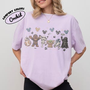 K&ouml;nnte beinhalten: Ein hellviolettes T-Shirt mit einem Grafikdruck von Star Wars-Figuren, darunter Chewbacca, ein Sturmtruppler, Grogu, R2-D2, Darth Vader und BB-8, die Luftballons halten. Der Text COMFORT COLORS Orchid steht in einem schwarzen Oval in der Ecke.