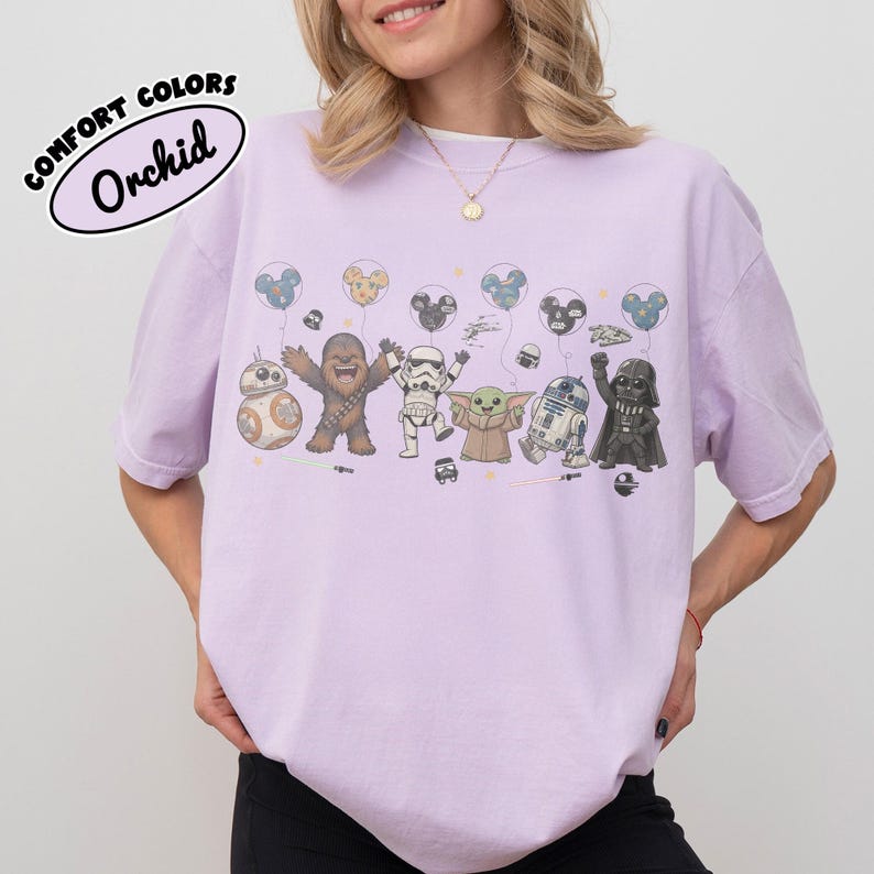 K&ouml;nnte beinhalten: Ein hellviolettes T-Shirt mit einem Grafikdruck von Star Wars-Figuren, darunter Chewbacca, ein Sturmtruppler, Grogu, R2-D2, Darth Vader und BB-8, die Luftballons halten. Der Text COMFORT COLORS Orchid steht in einem schwarzen Oval in der Ecke.