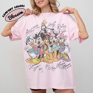 May include: A light pink t-shirt featuring a graphic of Disney characters including Mickey Mouse, Minnie Mouse, Donald Duck, Goofy, and Pluto. The shirt has the words "Comfort Colors" and "Blossom" in a circular design.