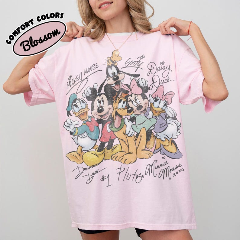 May include: A light pink t-shirt featuring a graphic of Disney characters including Mickey Mouse, Minnie Mouse, Donald Duck, Goofy, and Pluto. The shirt has the words "Comfort Colors" and "Blossom" in a circular design.