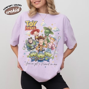 May include: A lilac Comfort Colors t-shirt with a Toy Story graphic. The design features Woody, Buzz Lightyear, and other characters, along with the phrase "You've got a Friend in me". The shirt also has the word "orchid" in a circle.