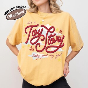 May include: A mustard yellow short-sleeve t-shirt featuring a graphic design with the red rope-style text "it's a Toy Story" and the script text "Baby, just say "yes"" accompanied by white cloud illustrations, small yellow stars, and red floral accents.