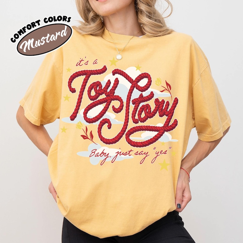 May include: A mustard yellow short-sleeve t-shirt featuring a graphic design with the red rope-style text "it's a Toy Story" and the script text "Baby, just say "yes"" accompanied by white cloud illustrations, small yellow stars, and red floral accents.