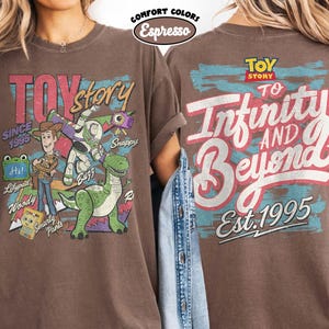 Pode incluir: Uma camiseta marrom de manga curta com gr&aacute;ficos de Toy Story. A frente mostra Woody, Buzz Lightyear, Rex e outros personagens com texto incluindo Toy Story, Since 1995 e nomes. As costas apresentam o texto To Infinity and Beyond.