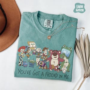 May include: A light green crew neck t-shirt featuring a graphic print of various animated characters including a cowboy, space ranger, cowgirl, bear, and alien. The text "YOU'VE GOT A FRIEND IN ME" is printed in black below the characters.