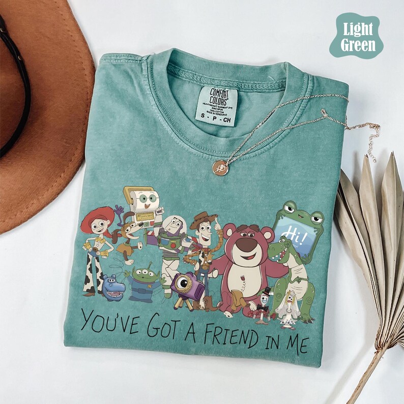 May include: A light green crew neck t-shirt featuring a graphic print of various animated characters including a cowboy, space ranger, cowgirl, bear, and alien. The text "YOU'VE GOT A FRIEND IN ME" is printed in black below the characters.