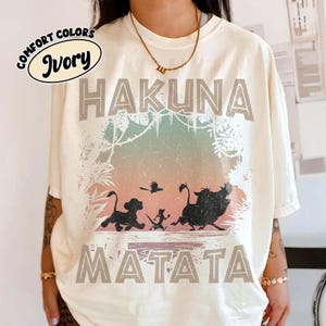 May include: Cream-coloured t-shirt with the words "HAKUNA MATATA" in grey block letters. The shirt features a colourful graphic of silhouettes of characters from the Lion King film. The words "Comfort Colors" and "Ivory" are in a circular design.
