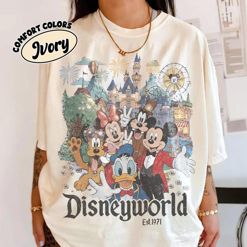 May include: Ivory-colored t-shirt featuring a colorful graphic of Disney characters, including Mickey Mouse, Minnie Mouse, and Donald Duck, in front of a castle and fireworks. The text "Disneyworld Est.1971" is printed below the graphic.