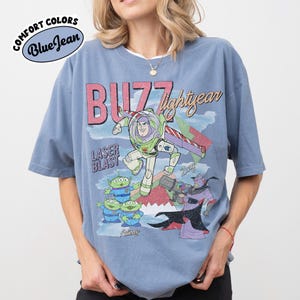 May include: A blue Comfort Colors t-shirt with a graphic of Buzz Lightyear and the text "Buzz Lightyear Laser Blast". The design includes aliens and Zurg. The shirt has short sleeves and a crew neck.