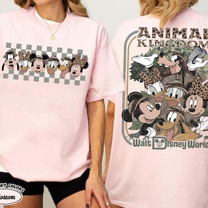 K&ouml;nnte beinhalten: Hellrosa T-Shirt mit einem Cartoon-Design von Disney-Figuren. Auf der Vorderseite befinden sich Gesichter von Figuren auf einem karierten Hintergrund. Auf der R&uuml;ckseite des Shirts stehen die Worte "ANIMAL KINGDOM" und "Walt Disney World".