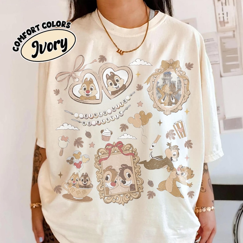 May include: Ivory-colored t-shirt featuring cartoon characters and the text "Disney with my bestie." The shirt has various framed images of cartoon characters, popcorn, and other graphics. The shirt is a Comfort Colors brand.