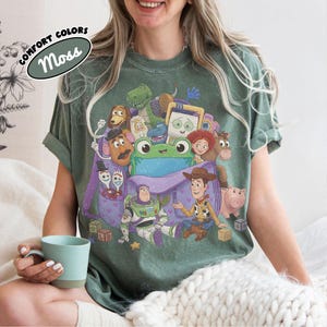 Pode incluir: Uma t-shirt verde-musgo com uma estampa gr&aacute;fica de v&aacute;rios personagens animados da franquia Toy Story, incluindo Woody, Buzz Lightyear e um sapo verde central, todos reunidos em torno de uma mochila roxa com padr&otilde;es de cora&ccedil;&otilde;es.