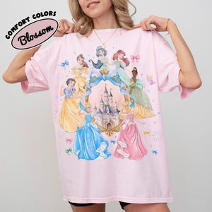May include: A light pink t-shirt featuring a design of several cartoon princesses surrounding a castle. The shirt has the words "Comfort Colors" and "Blossom" printed on the top left corner.