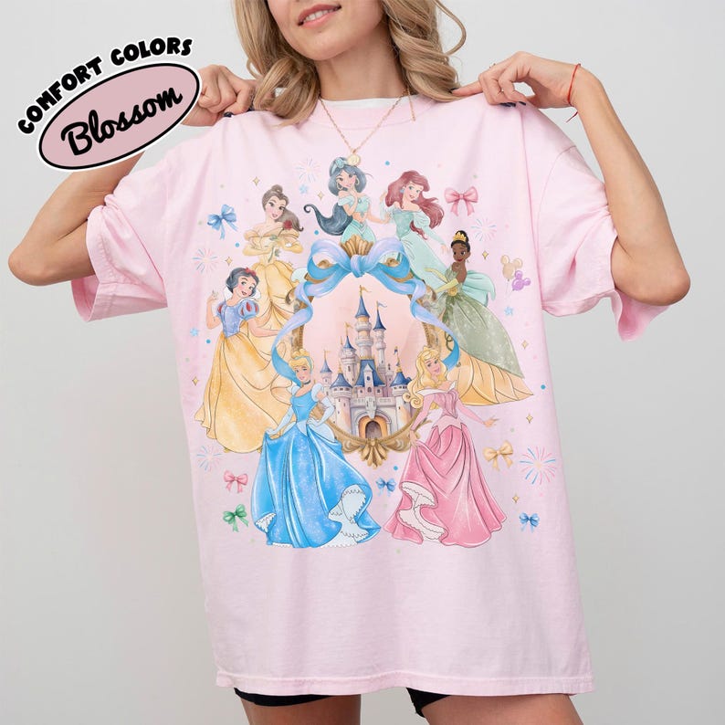 May include: A light pink t-shirt featuring a design of several cartoon princesses surrounding a castle. The shirt has the words "Comfort Colors" and "Blossom" printed on the top left corner.