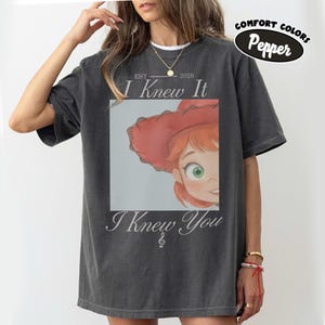 Op de afbeelding: Een antracietgrijs T-shirt met korte mouwen en een grafische print van een stripfiguur met rood haar en een rode hoed. De tekst luidt EST 2026 I Knew It I Knew You met een vioolsleutelsymbool. Een klein ovaal label toont Comfort Colors Pepper.