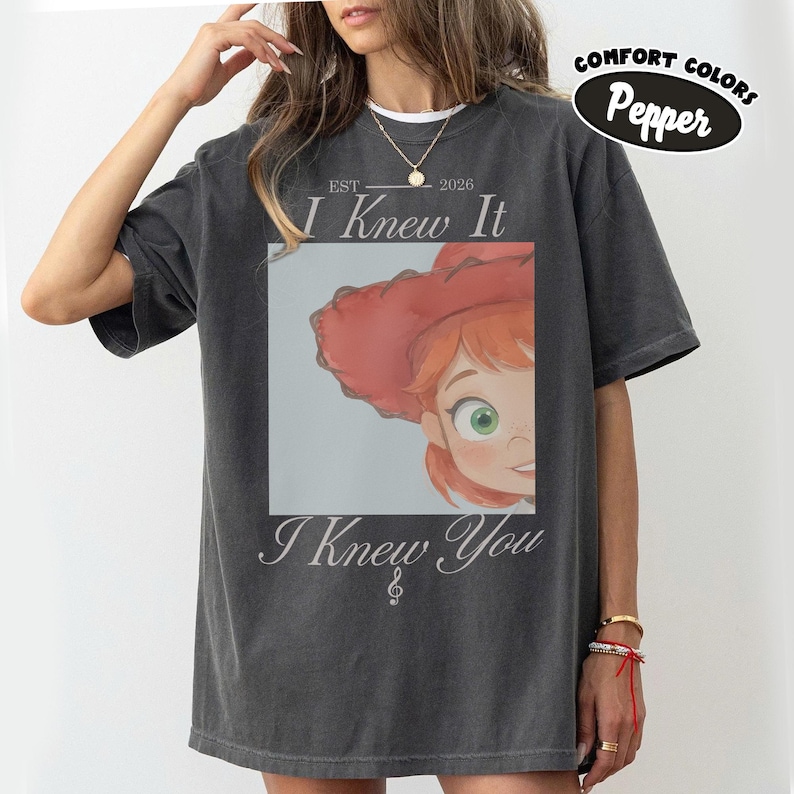 Op de afbeelding: Een antracietgrijs T-shirt met korte mouwen en een grafische print van een stripfiguur met rood haar en een rode hoed. De tekst luidt EST 2026 I Knew It I Knew You met een vioolsleutelsymbool. Een klein ovaal label toont Comfort Colors Pepper.