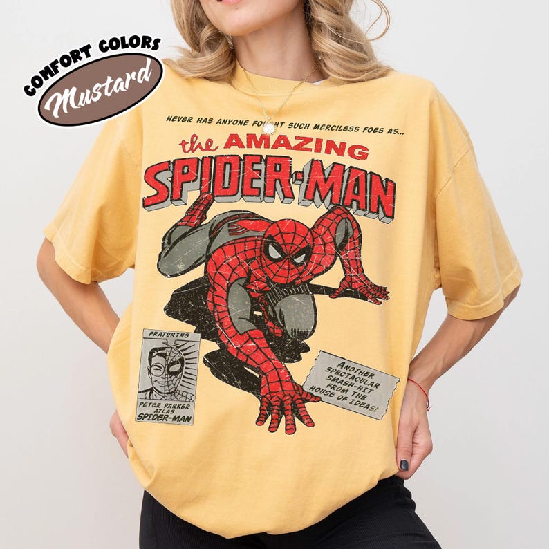 May include: A mustard yellow short-sleeve t-shirt featuring a distressed graphic of Spider-Man. The text reads: NEVER HAS ANYONE FOUGHT SUCH MERCILESS FOES AS... the AMAZING SPIDER-MAN, FRATURING PETER PARKER ATLAS SPIDER-MAN, and ANOTHER SPECTACULAR SMASH-HIT FROM THE HOUSE OF IDEAS!