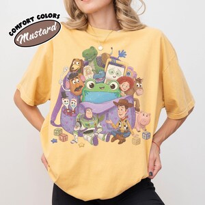 Pode incluir: Uma t-shirt amarelo-mostarda com uma estampa gr&aacute;fica de v&aacute;rios personagens animados da franquia Toy Story saindo de uma mochila roxa, com o texto COMFORT COLORS MUSTARD impresso no canto superior esquerdo da imagem.
