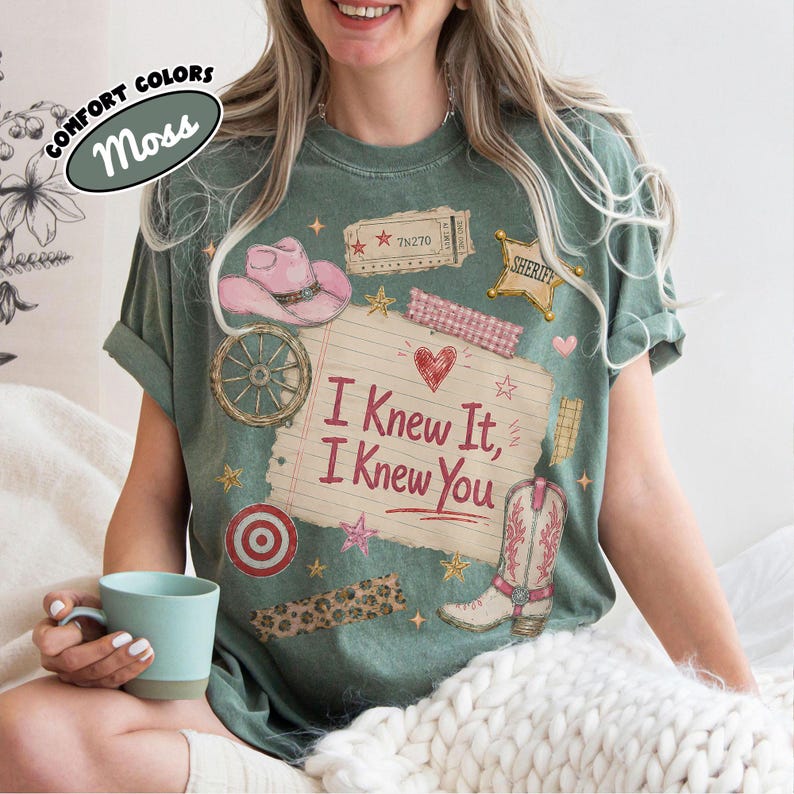 Puede incluir: Una camiseta verde musgo con un estampado gr&aacute;fico de una p&aacute;gina de cuaderno rayada con el texto I Knew It, I Knew You. El dise&ntilde;o incluye un sombrero de vaquero rosa, una estrella de sheriff, una rueda de carreta, una diana, una bota y un boleto.