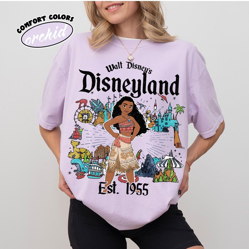 Op de afbeelding: Een lichtpaars t-shirt met korte mouwen en een grafische print van Moana in het midden, omringd door diverse Disneyland-parkiconen. De tekst luidt Walt Disney's Disneyland Est. 1955. Een klein ovaal logo linksboven vermeldt Comfort Colors orchid.