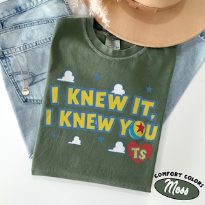 May include: An olive green t-shirt featuring the yellow text I KNEW IT, I KNEW YOU, a colorful ball graphic with a red star, small white cloud shapes, blue stars, and a red heart containing the yellow letters TS, with a tag reading XL-TG-EG.