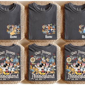 Peut inclure: Six t-shirts gris fonc&eacute; avec des graphiques sur le th&egrave;me de Disney. La rang&eacute;e sup&eacute;rieure pr&eacute;sente des motifs de personnages avec le mot "Name" en dessous. La rang&eacute;e inf&eacute;rieure affiche un motif Disneyland avec Mickey Mouse et ses amis, les mots "Walt Disney's Disneyland Est. 1955".