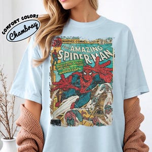 May include: A light blue t-shirt featuring a vintage Marvel Comics The Amazing Spider-Man graphic print on the front. The design includes Spider-Man in a dynamic pose with comic book text and a distressed, weathered texture throughout the image.
