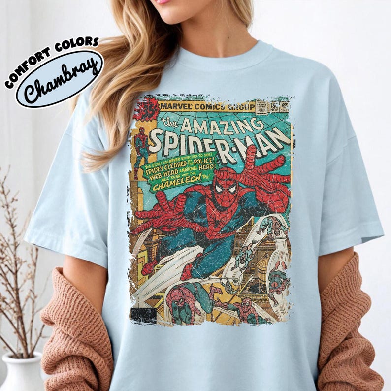 May include: A light blue t-shirt featuring a vintage Marvel Comics The Amazing Spider-Man graphic print on the front. The design includes Spider-Man in a dynamic pose with comic book text and a distressed, weathered texture throughout the image.