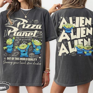 May include: Dark grey t-shirt featuring the Pizza Planet logo and three green aliens. The front of the shirt has text that reads "Pizza Planet" and "Out of this world quality". The back of the shirt has the word "ALIEN" repeated three times.