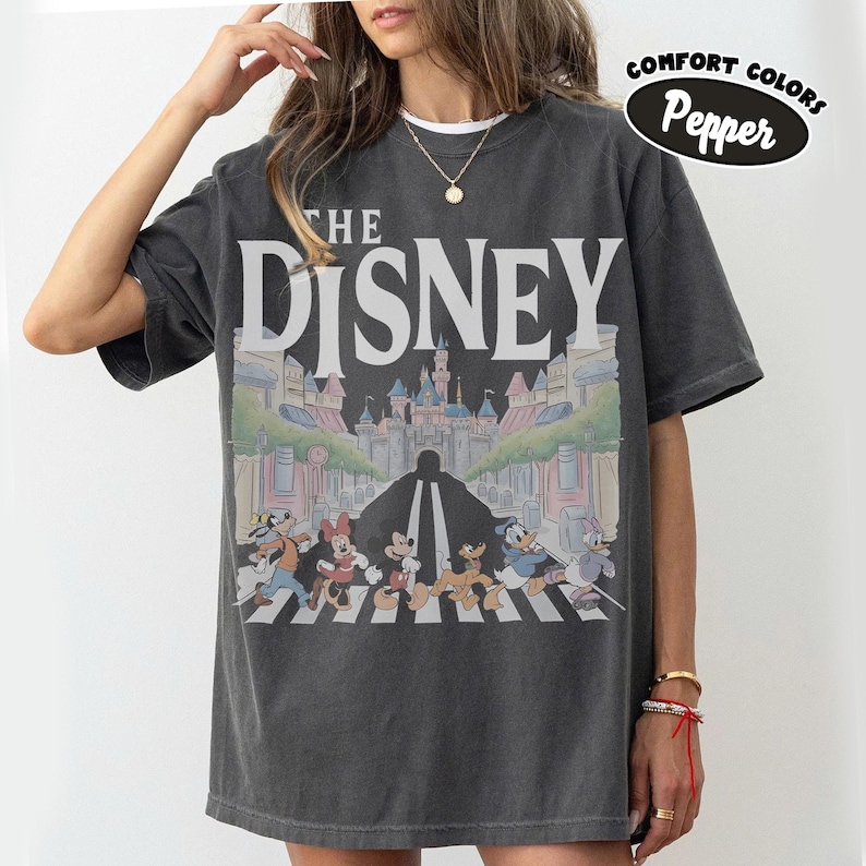 Może przedstawiać: Ciemnoszara koszulka z napisem "THE DISNEY" dużymi białymi literami. Grafika przedstawia postacie Disneya przechodzące przez ulicę, z zamkiem w tle. Koszulka ma wygląd vintage, a słowa "Comfort Colors Pepper" są w czarnym owalu.