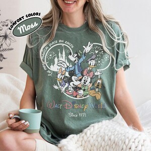 May include: Moss green t-shirt featuring a graphic of Disney characters including Mickey Mouse, Minnie Mouse, Donald Duck, and Goofy. The shirt has the text "Walt Disney World Since 1971" and "The Most Magical Place on Earth".