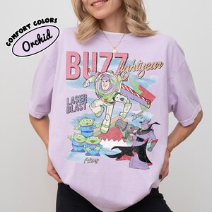 May include: A lilac t-shirt with a graphic of Buzz Lightyear, aliens, and Zurg from Toy Story. The shirt has the words "Buzz Lightyear" and "Laser Blast" printed on it. The shirt is labelled "Comfort Colors" and "Orchid".