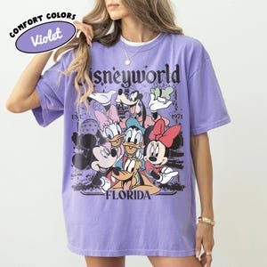 May include: A violet Comfort Colors t-shirt featuring a Disneyworld graphic with Mickey Mouse, Minnie Mouse, Donald Duck, Daisy Duck, Goofy, and Pluto. The shirt has the text "Disneyworld Florida" and "Est. 1971".