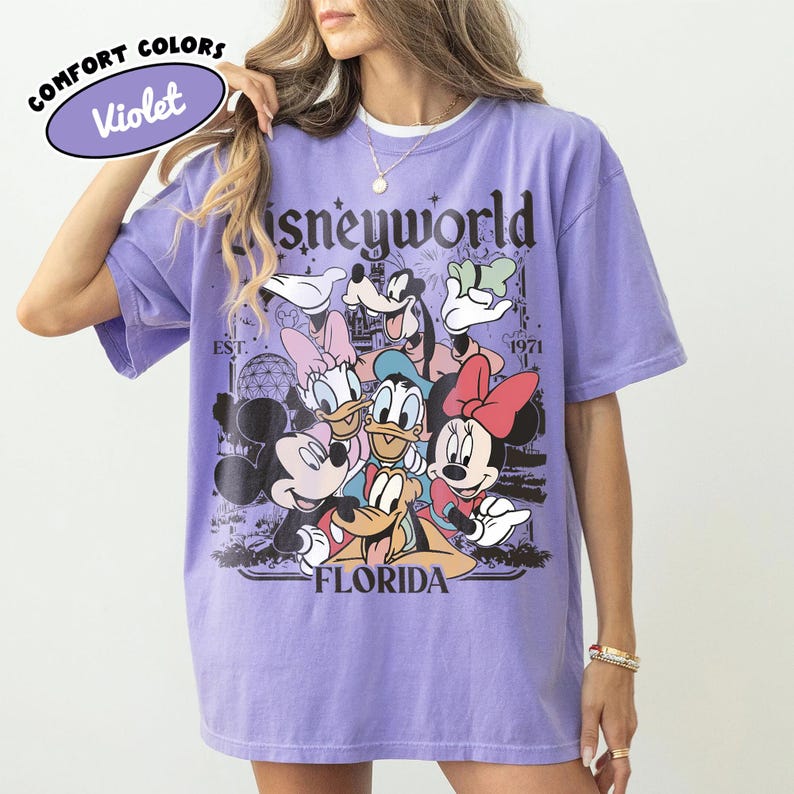 May include: A violet Comfort Colors t-shirt featuring a Disneyworld graphic with Mickey Mouse, Minnie Mouse, Donald Duck, Daisy Duck, Goofy, and Pluto. The shirt has the text "Disneyworld Florida" and "Est. 1971".