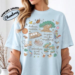 Puede incluir: Camiseta azul claro con un dise&ntilde;o gr&aacute;fico colorido que presenta personajes de dibujos animados, un &aacute;rbol y las frases "hakuna matata" y "adventure is out there". La camiseta tiene las palabras "COMFORT COLORS" y "Chambray" en un c&iacute;rculo.