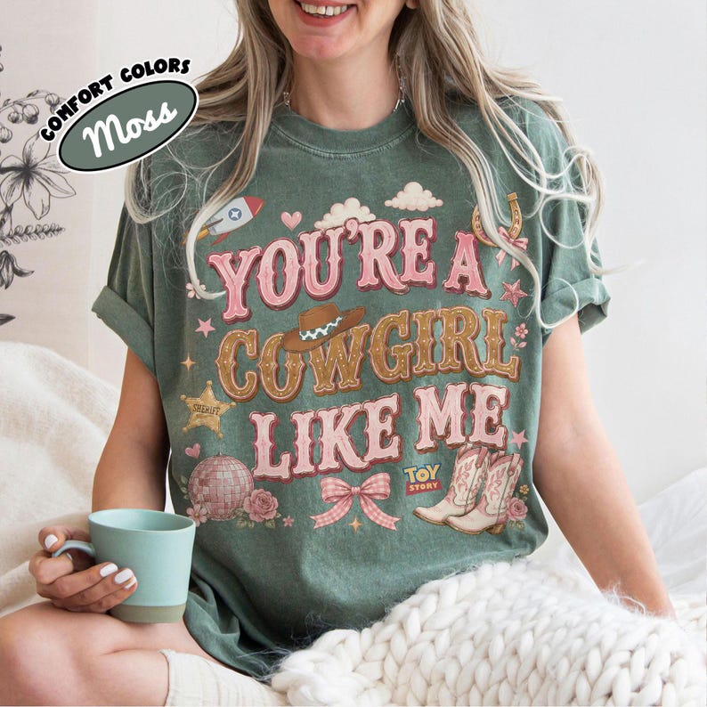 Puede incluir: Una camiseta de manga corta color verde musgo con el texto "YOU'RE A COWGIRL LIKE ME" en rosa y marr&oacute;n. El gr&aacute;fico incluye un sombrero de vaquero, un cohete, una estrella de sheriff, una bola de discoteca, un lazo y botas con "TOY STORY".