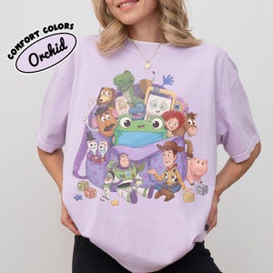 Pode incluir: Uma t-shirt roxo-claro com uma estampa gr&aacute;fica de v&aacute;rios personagens de brinquedos animados reunidos em torno de uma mochila roxa. O design inclui um sapo verde, um cowboy, um patrulheiro espacial e outras figuras com blocos na base.