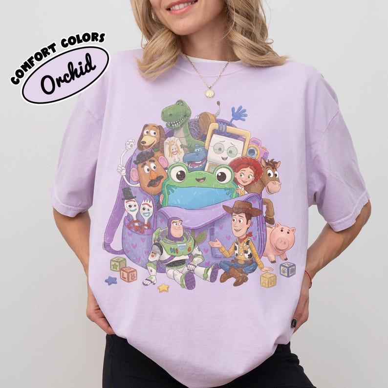 Pode incluir: Uma t-shirt roxo-claro com uma estampa gr&aacute;fica de v&aacute;rios personagens de brinquedos animados reunidos em torno de uma mochila roxa. O design inclui um sapo verde, um cowboy, um patrulheiro espacial e outras figuras com blocos na base.