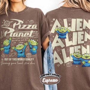 May include: Brown t-shirt with the Pizza Planet logo and three green aliens in blue suits. The text reads "The Claw chooses who will go and who will stay." The back of the shirt says "Alien" multiple times.