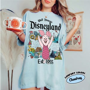 Op de afbeelding: Een lichtblauw t-shirt met een afbeelding van Knorretje uit Winnie de Poeh, gecentreerd boven de tekst Walt Disney's Disneyland Est. 1955. Het ontwerp bevat diverse parkiconen zoals een kasteel, een trein en een raket in gekleurde inkt.