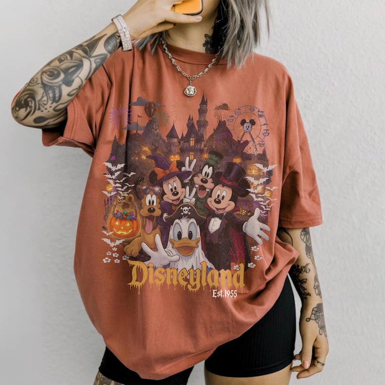 May include: A rust-colored short-sleeve t-shirt featuring a graphic print of Disney characters in Halloween costumes, including Mickey Mouse, Minnie Mouse, Donald Duck, Goofy, and Pluto. The text reads Disneyland Est. 1955 with a castle background.
