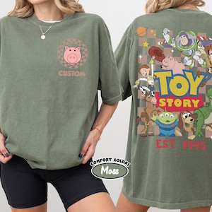 Puede incluir: Camiseta verde oliva con un gr&aacute;fico de cerdo rosa y la palabra "CUSTOM" en la parte delantera. La parte trasera de la camiseta muestra un gr&aacute;fico colorido de "Toy Story" con varios personajes y el texto "EST. 1995".