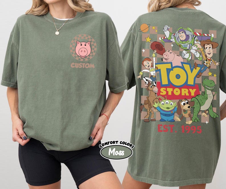 Puede incluir: Camiseta verde oliva con un gr&aacute;fico de cerdo rosa y la palabra "CUSTOM" en la parte delantera. La parte trasera de la camiseta muestra un gr&aacute;fico colorido de "Toy Story" con varios personajes y el texto "EST. 1995".