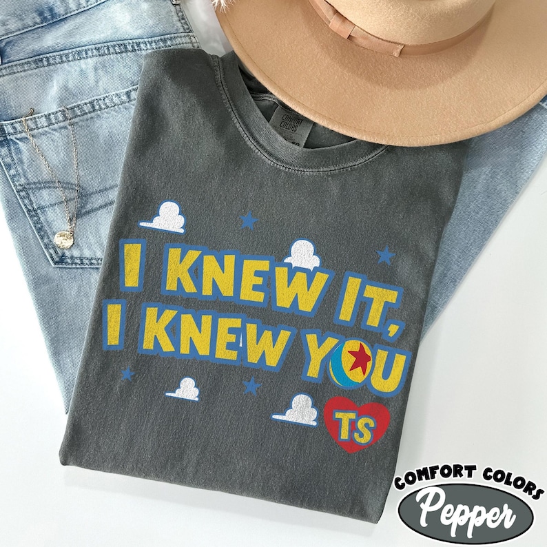 May include: A charcoal gray crew neck t-shirt featuring the yellow text I KNEW IT, I KNEW YOU with a colorful ball graphic, small white cloud icons, blue stars, and a red heart containing the yellow letters TS printed on the front.